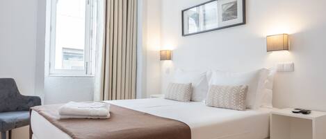 A modern hotel room with a large bed, bedside tables, a chair, and a window with curtains.