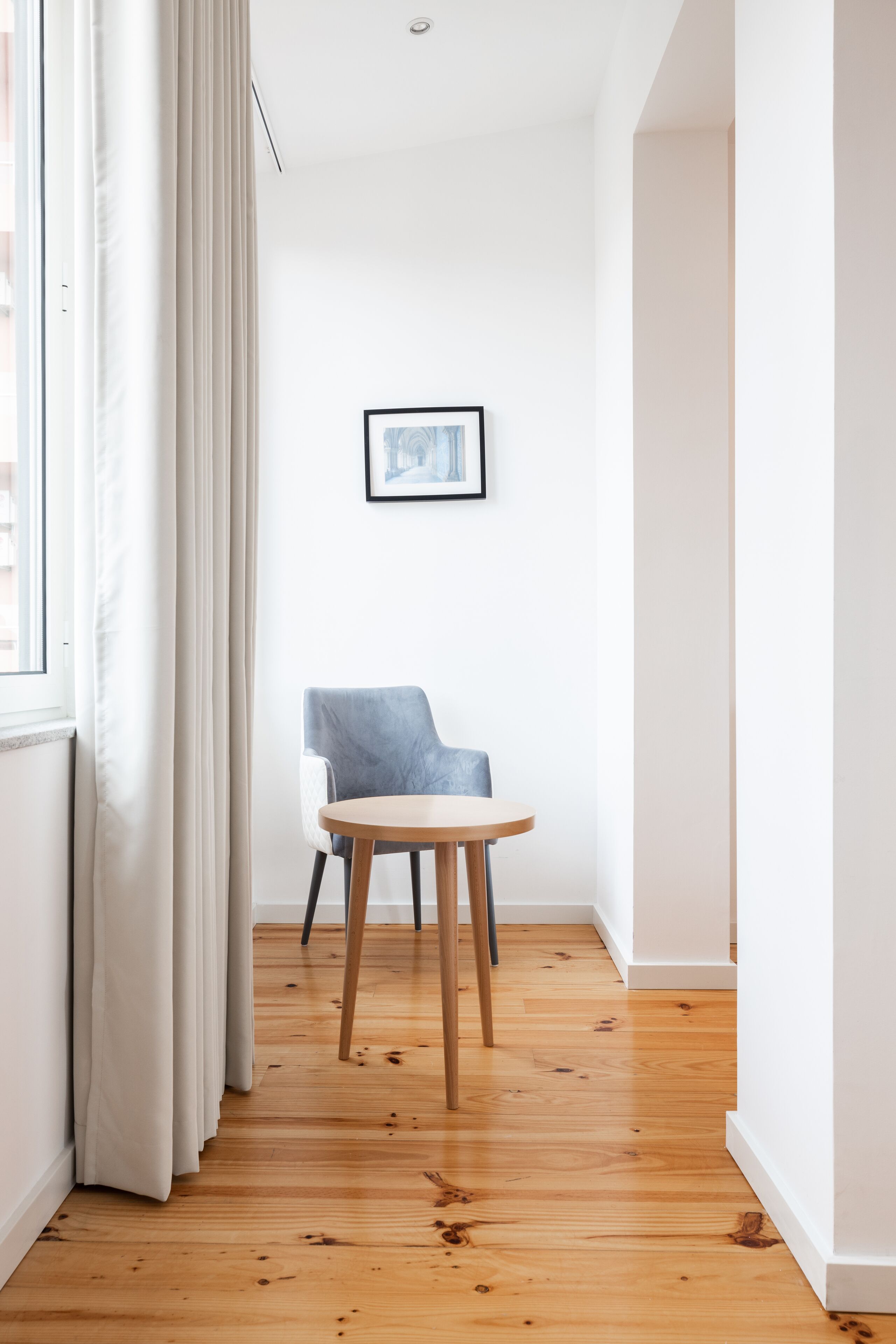 A hallway with wooden flooring, a blue chair, a small round table, and a framed picture on the wall.