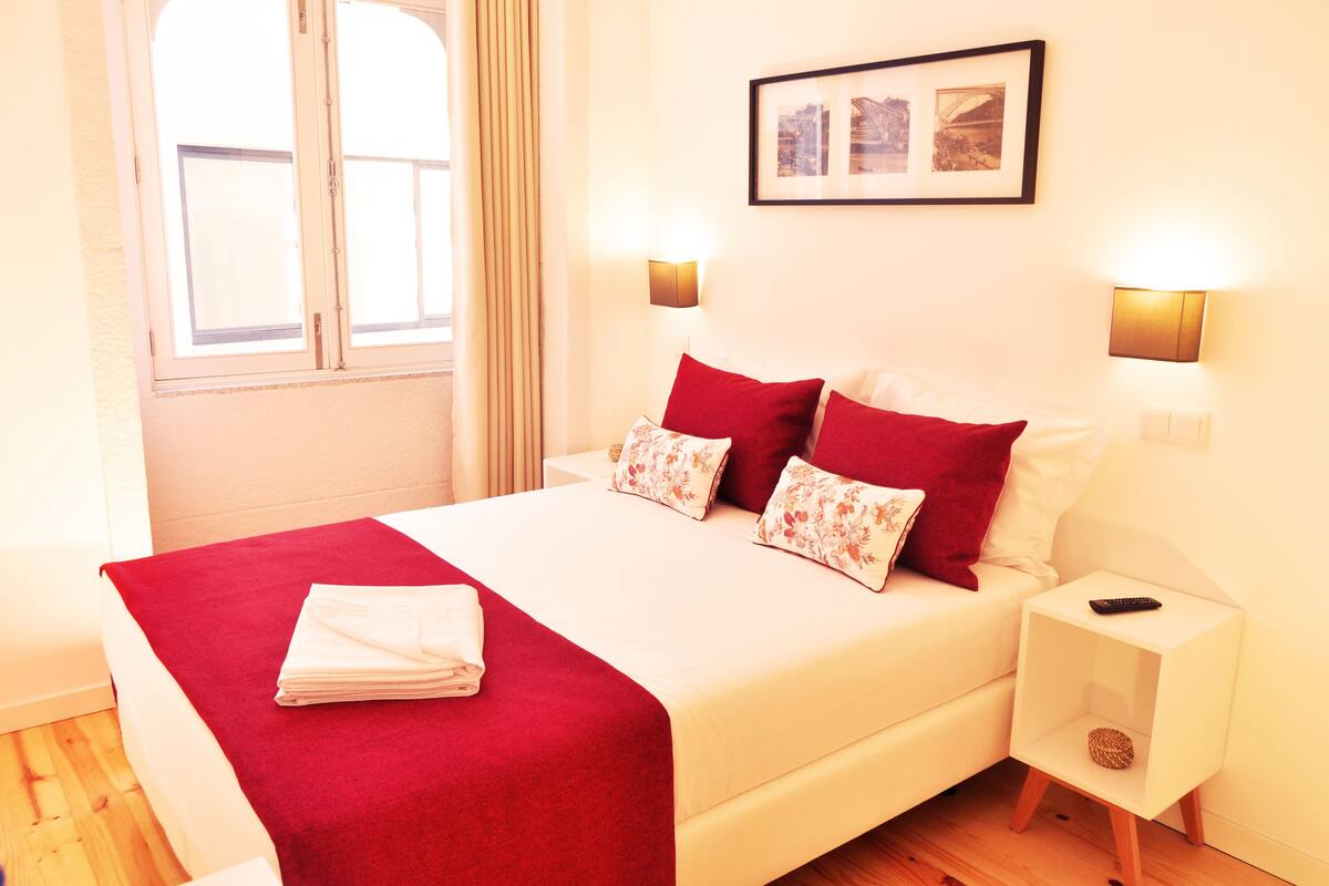 A neatly made bed with red and white bedding, a small bedside table with a remote control, and a framed picture on the wall.