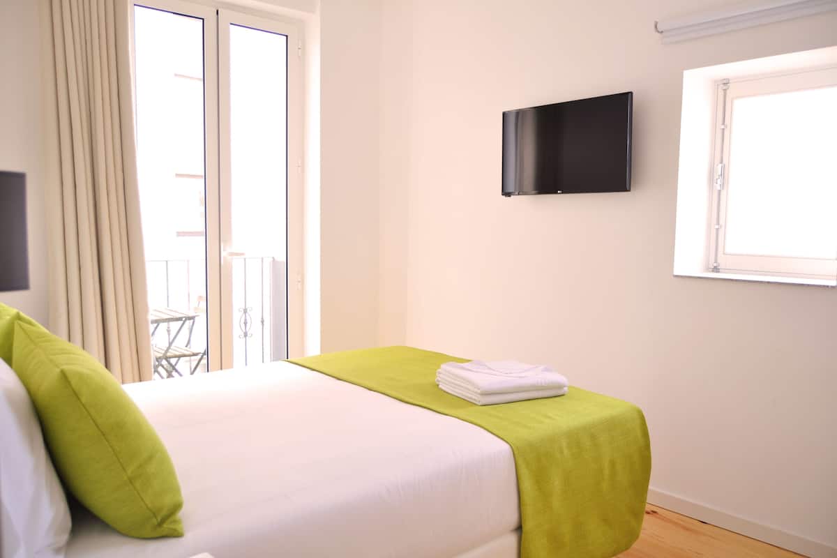 A hotel room with a bed, a green bedspread, a folded white towel, a TV mounted on the wall, and a window with curtains.