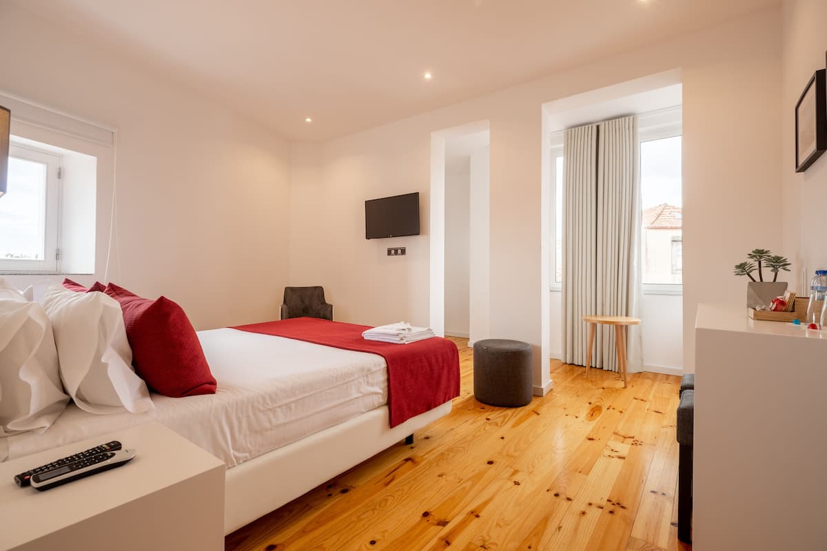 A modern hotel room with a wooden floor, a bed with white and red bedding, a TV mounted on the wall, and a small table with a plant.