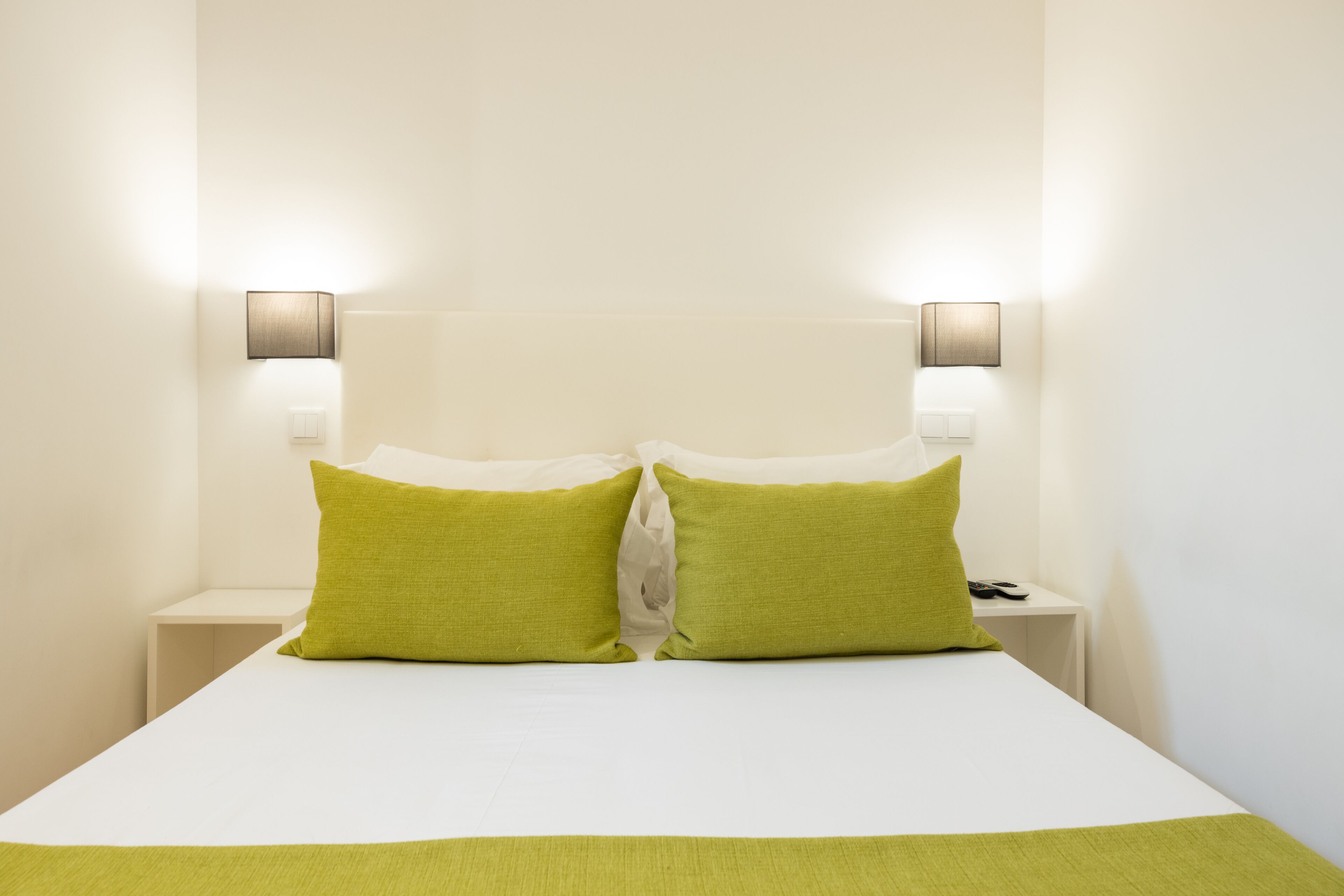 A neatly made bed with two pillows, a bedside table with a lamp, and a wall-mounted light fixture.