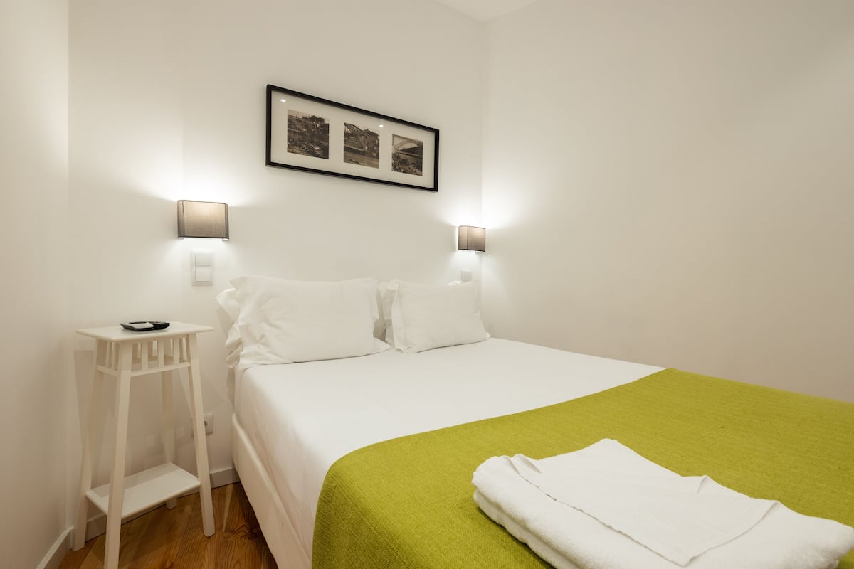 A neatly made bed with white linens and a green throw, two bedside lamps, and a small white nightstand with a lamp.