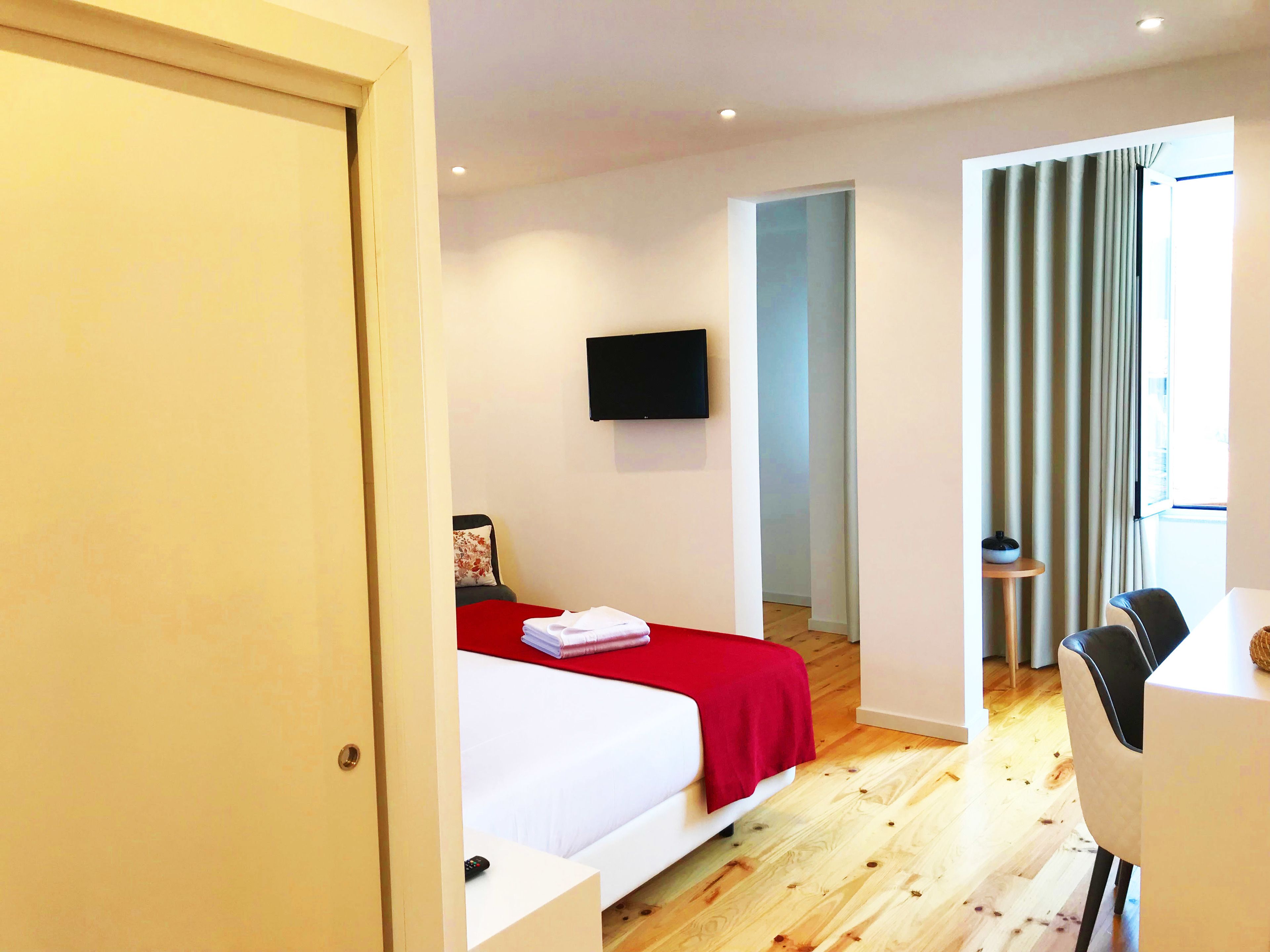 A modern hotel room with a bed, a TV mounted on the wall, and a dining area with a table and chairs.