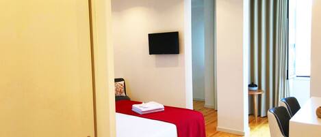 A modern hotel room with a bed, a TV mounted on the wall, and a dining area with a table and chairs.