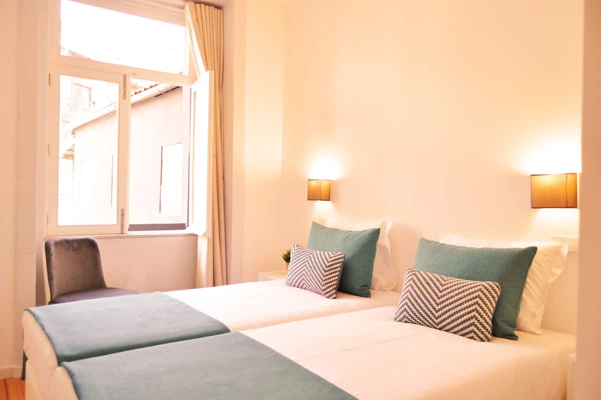 A hotel room with two beds, a chair, a window, and wall-mounted lights.