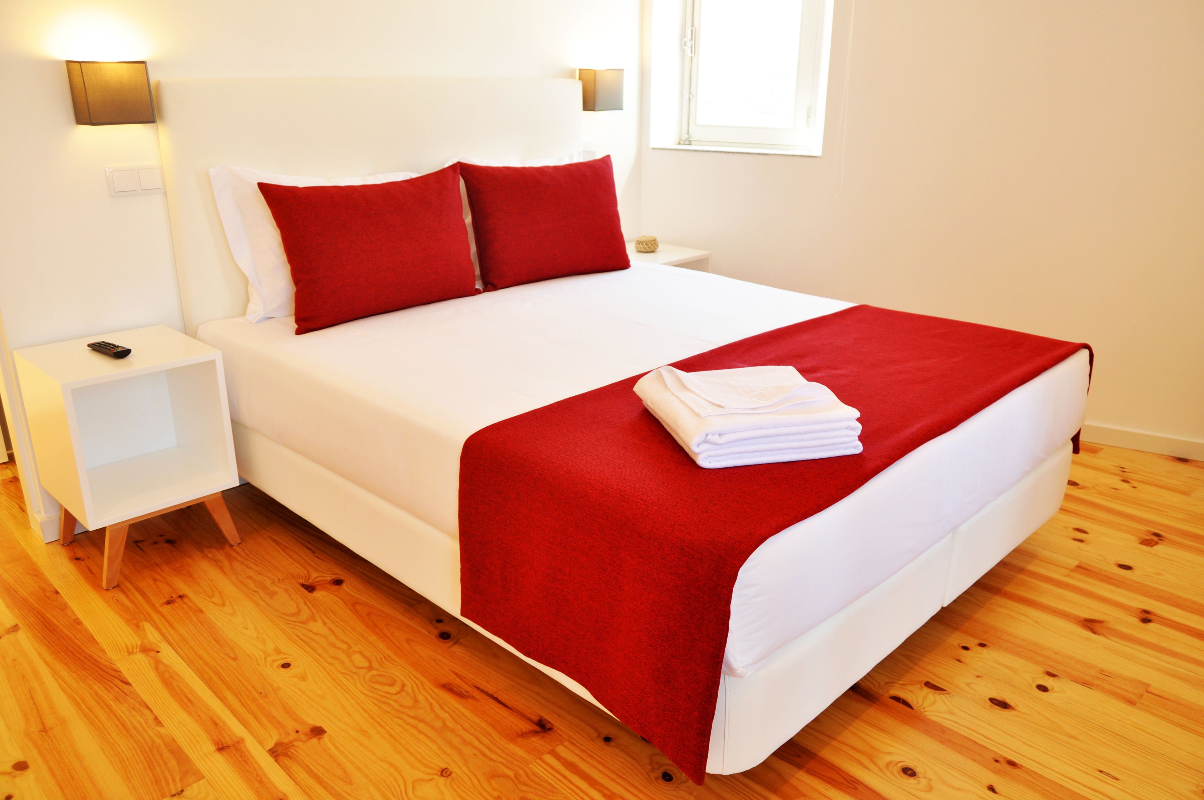 Un lit bien fait, avec une literie rouge et blanche, une table de chevet blanche avec une télécommande, et un plancher en bois.