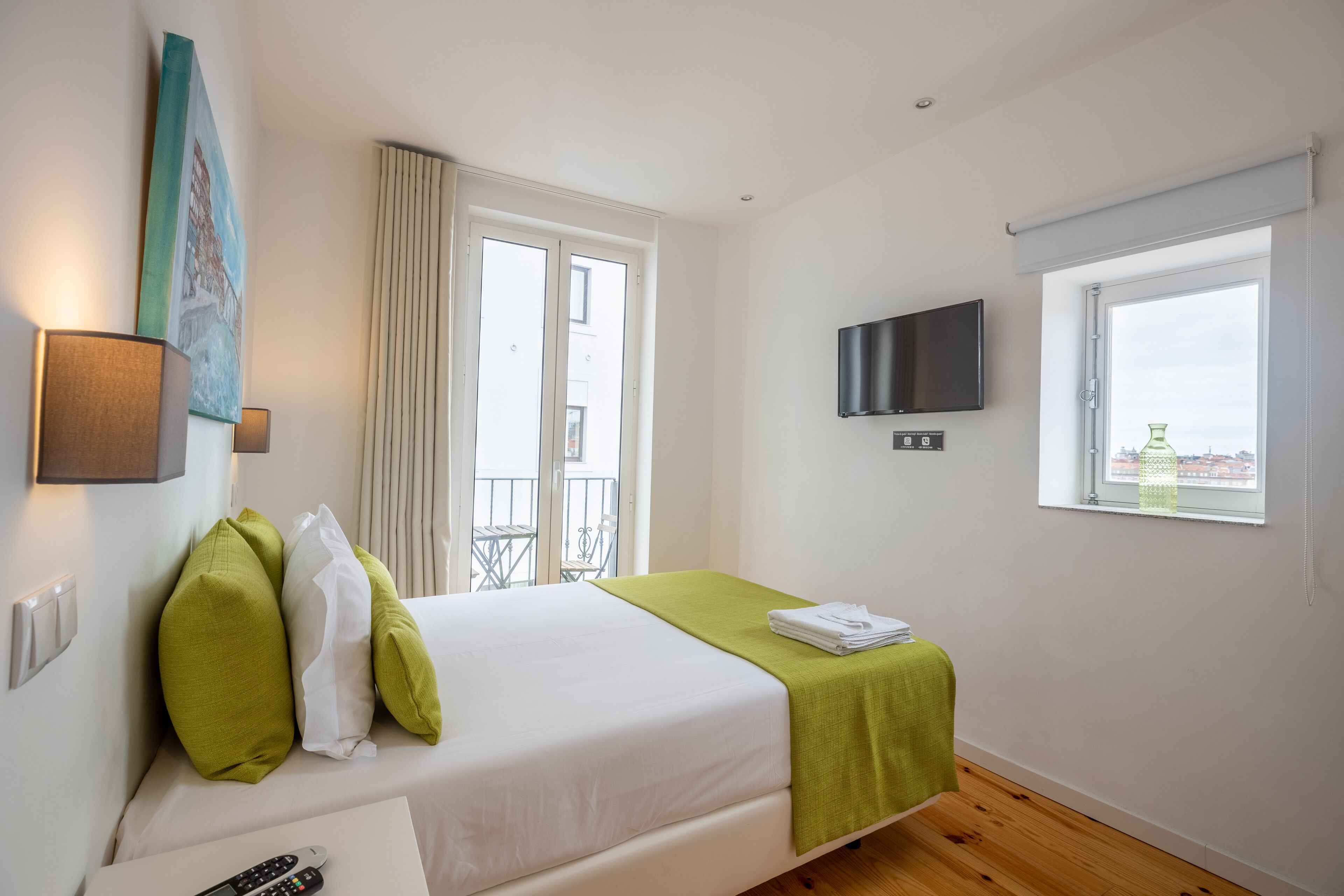 Habitación de hotel con cama, televisor montado en la pared, ventana con vistas y balcón con mesa y sillas.