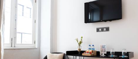 Una habitación moderna con piso de madera, una silla negra con cojín, una mesita con un florero y botellas, y un televisor montado en la pared.