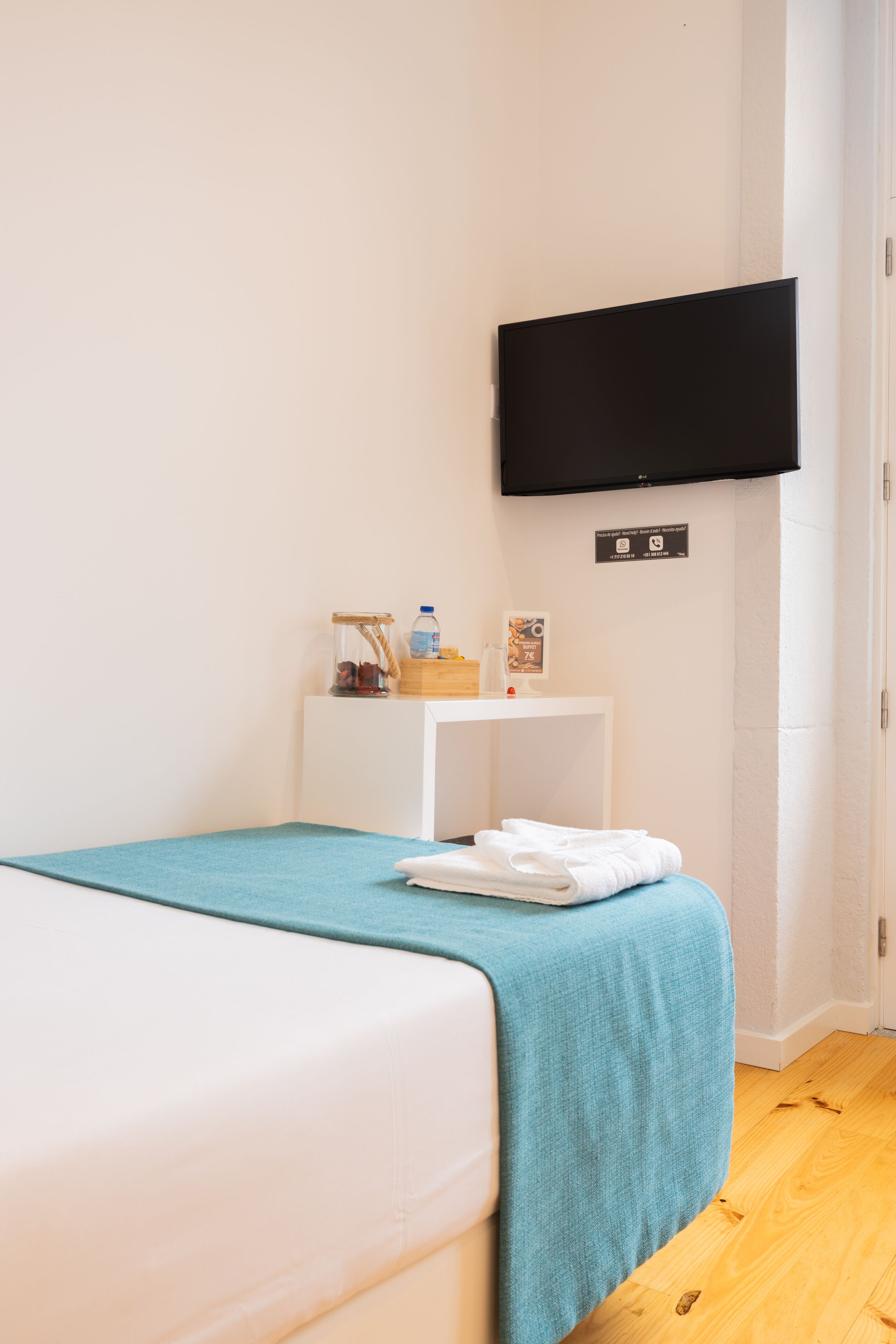 A hotel room with a bed, a small bedside table with toiletries, a flat-screen TV mounted on the wall, and a wooden floor.