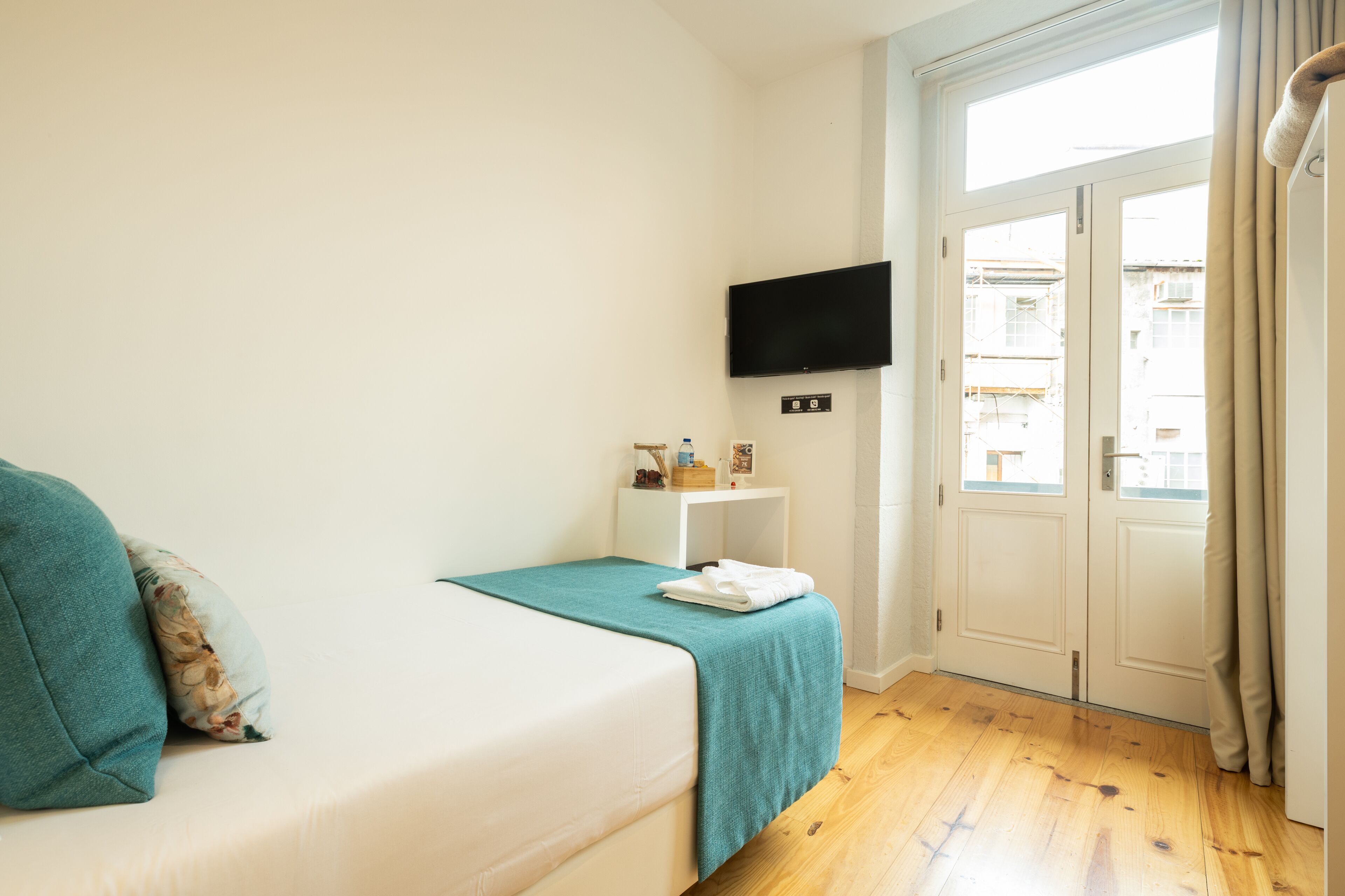 A bedroom with a bed, a TV mounted on the wall, a small bedside table with a bottle, and a window with curtains.