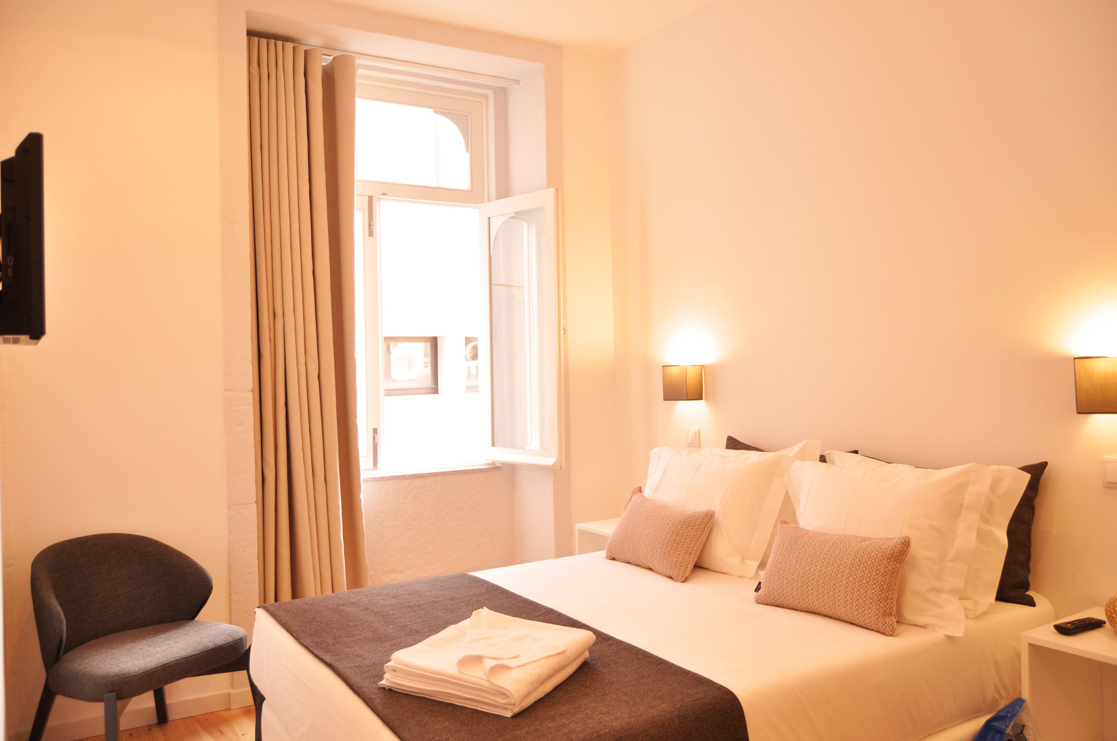Habitación de hotel con cama, una silla, una mesita de noche y un ventanal con cortinas.