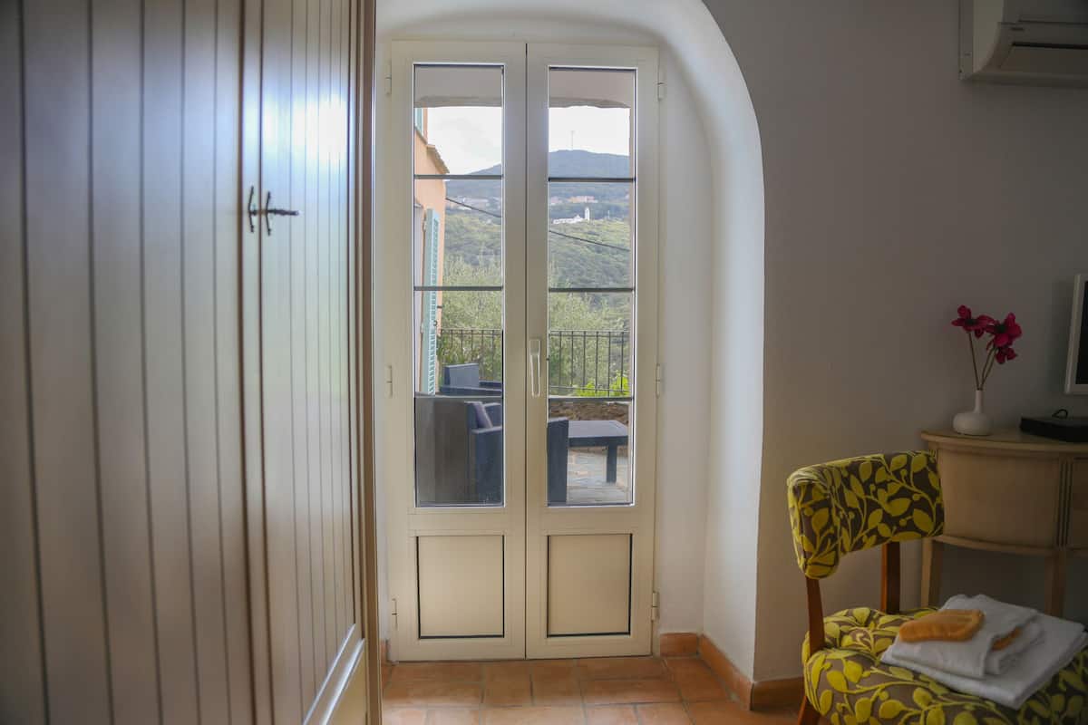 Een kamer met een schuifdeur die leidt naar een balkon, een stoel met een gekleurde kussen en een klein tafeltje met een vaas.