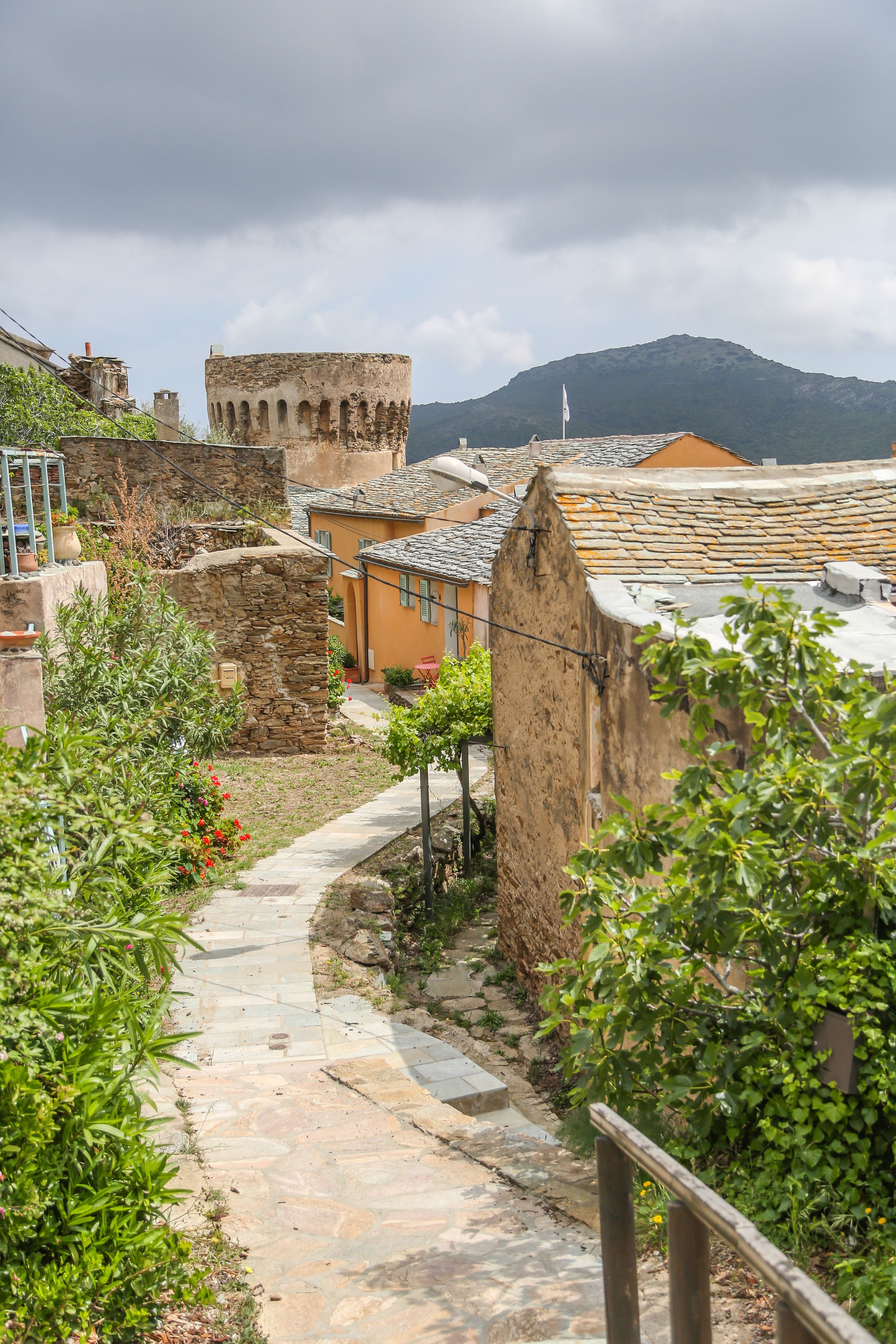 A stone pathway leading through a rustic village with traditional stone buildings and a tower in the background.