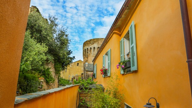 Une ruelle étroite, avec un bâtiment jaune, des volets verts et une tour de pierre en arrière-plan.