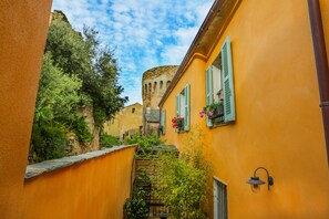 Une ruelle étroite, avec un bâtiment jaune, des volets verts et une tour de pierre en arrière-plan.