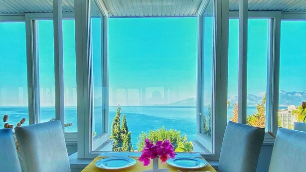 A dining table with yellow tablecloth, two white cups, and a vase of purple flowers, set against a backdrop of a scenic view through large windows. - Royal City Hotel (Antalya)
