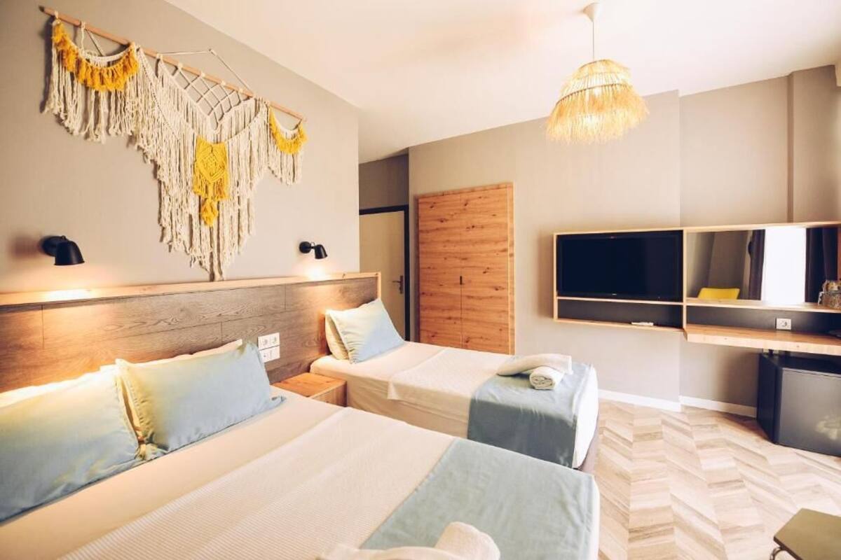 Quarto de hotel com duas camas, um guarda-roupa de madeira, televisão e um painel de parede em macramé.