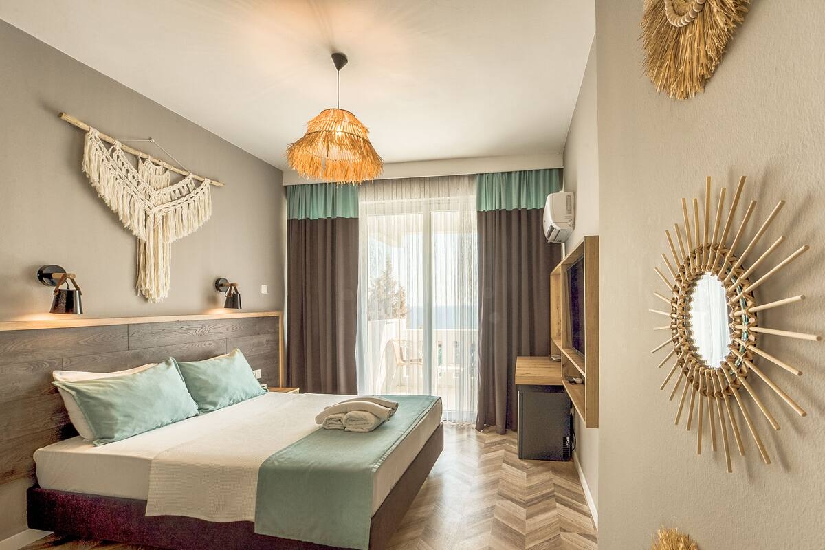 Quarto de hotel moderno com uma cama, decoração de parede em macramé pendente, ventilador de teto e um espelho grande com moldura em formato de sol.