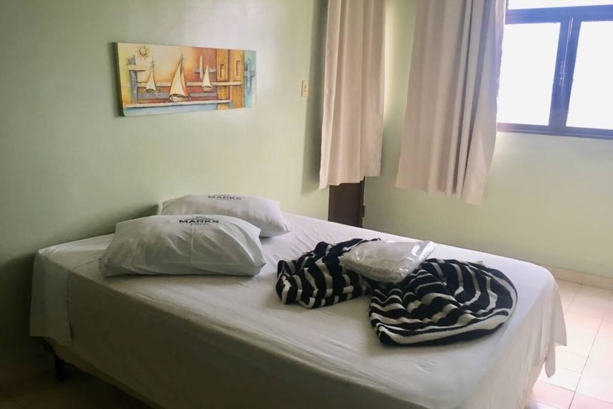 A single bed with white bedding and two pillows, a painting of sailboats on the wall, and a window with curtains.