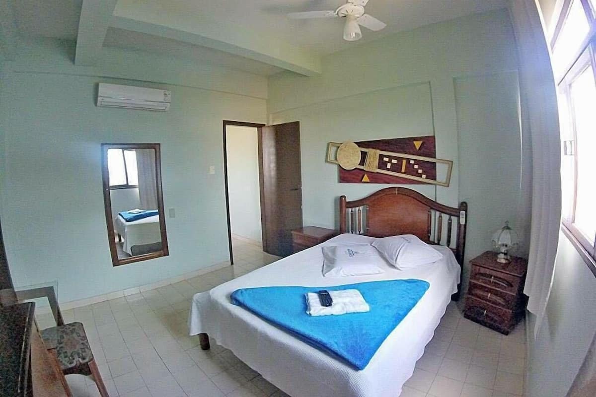 A hotel room with a bed, a ceiling fan, a mirror, a guitar decoration, and a window.