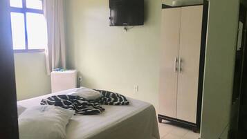 Quarto com cama, televisão fixada na parede, guarda-roupa e janela com cortinas.