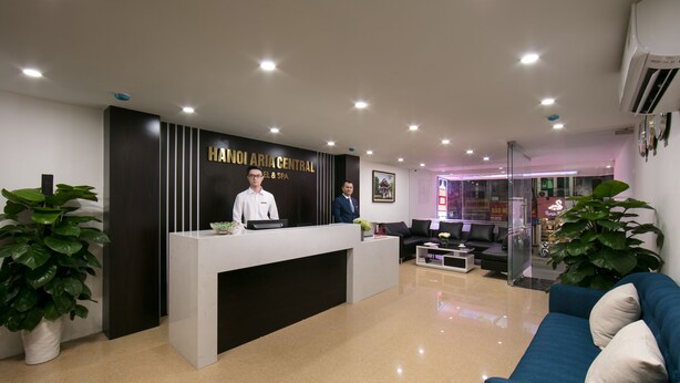 Zona de recepción de hotel con mostrador, dos empleados, un sofá azul y plantas en macetas.