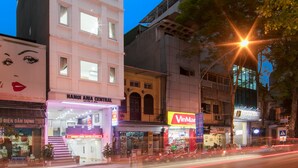 Un edificio de varios pisos con una aguja destacada, un letrero del Hotel Hanoi Amica Central y diversas tiendas, incluyendo VinMar.