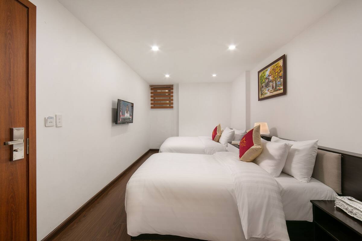 A hotel room with two beds, a wooden door, a painting on the wall, and a window with blinds.
