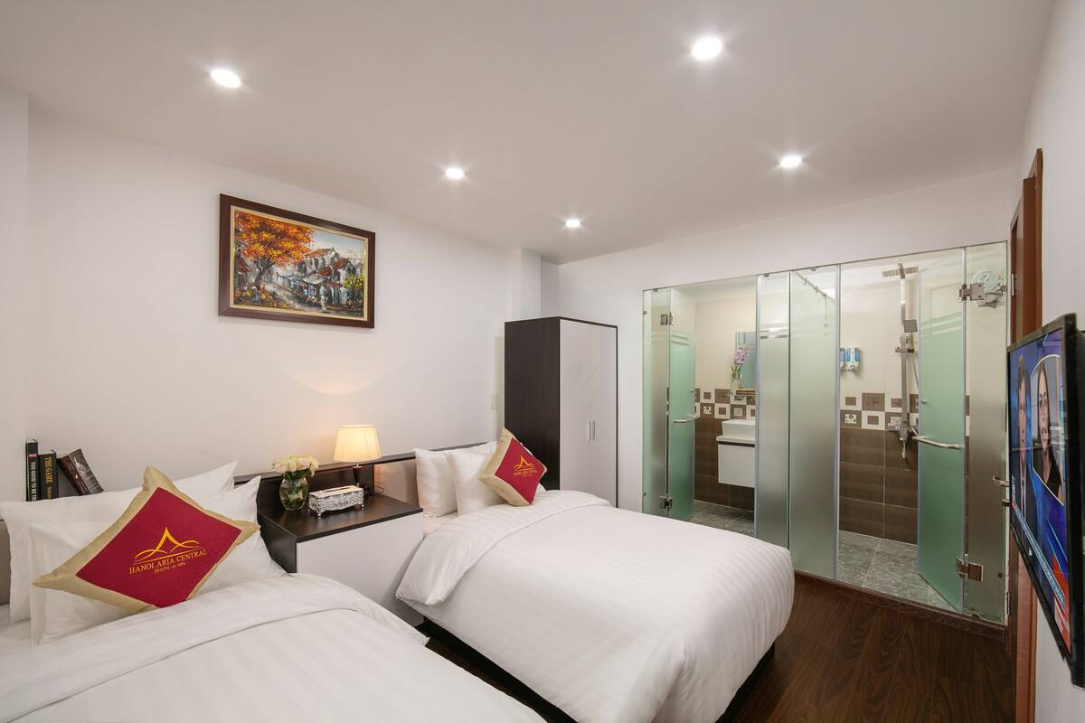 A hotel room with two beds, a painting on the wall, a nightstand with a lamp, and a glass-enclosed shower area.