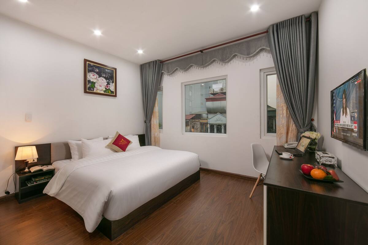 A hotel room with a bed, a desk with a laptop, a chair, a nightstand with a lamp, and a window with curtains.