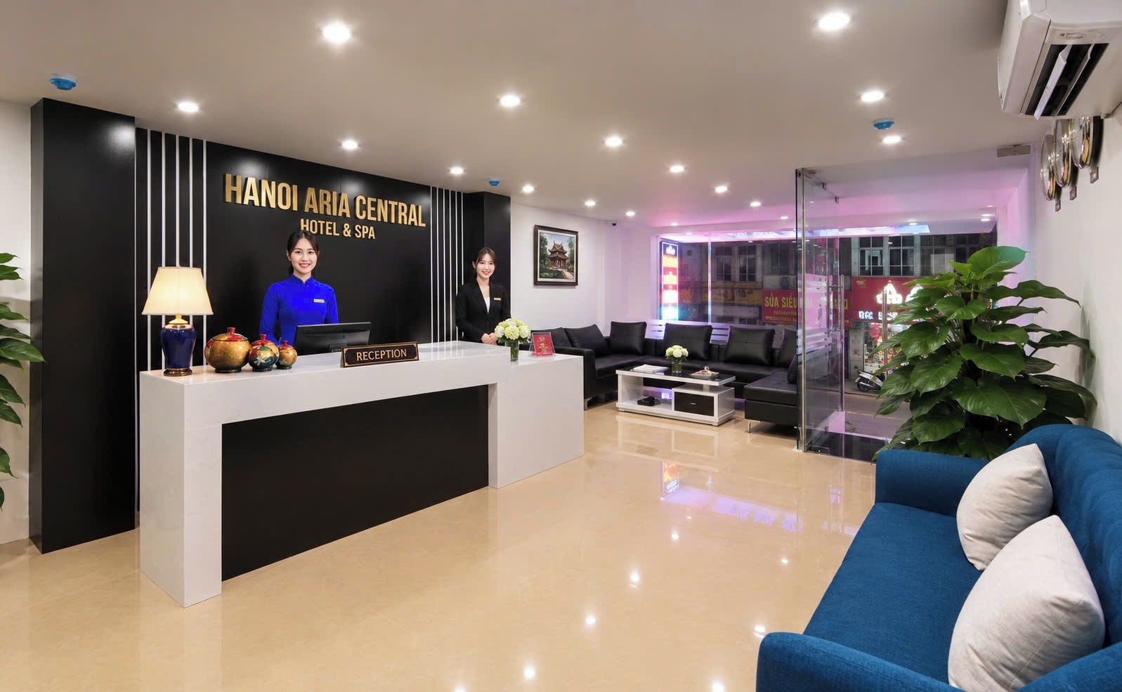A hotel reception area with a reception desk, two staff members, a sofa, and a large plant.