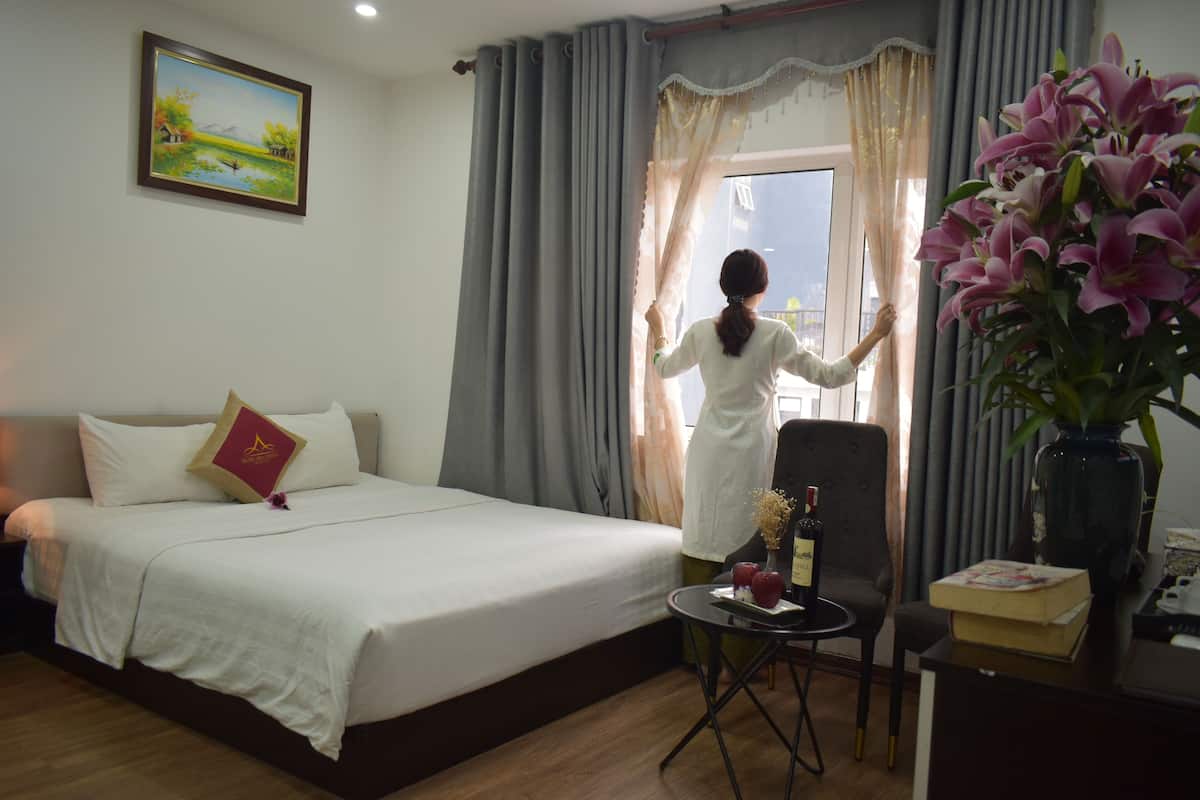 A hotel room with a bed, a chair, a small table with a bottle and glasses, a vase with flowers, and a painting on the wall.