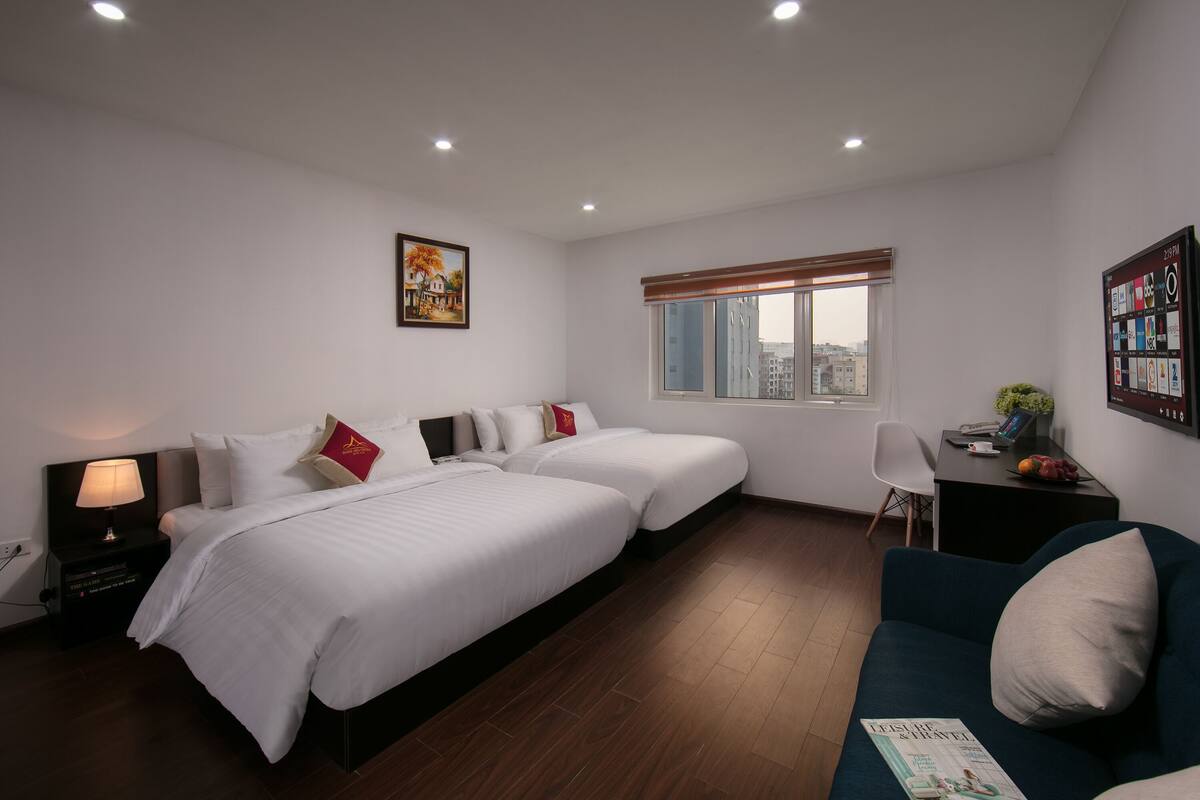 A hotel room with two beds, a desk, a chair, a window with a view of buildings, and a magazine on the sofa.