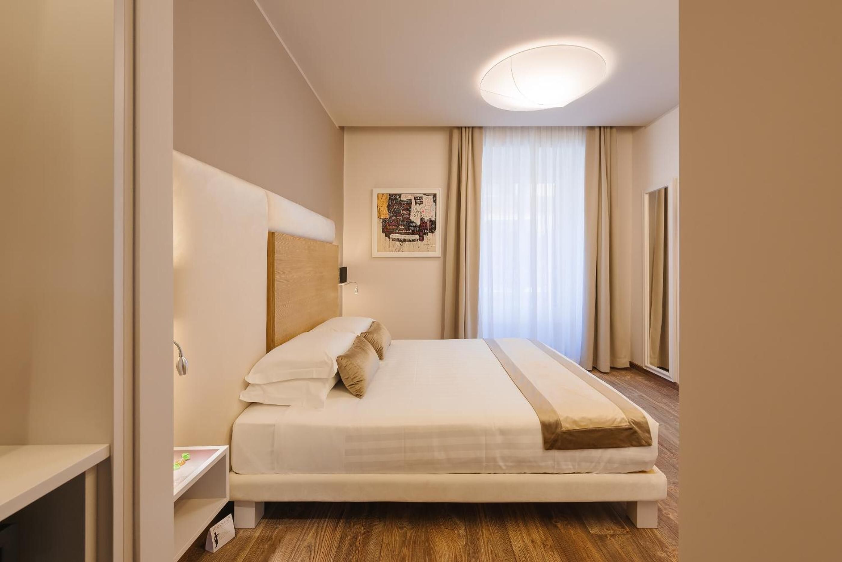 A neatly arranged hotel room with a large bed, a bedside table, a headboard, a wall-mounted artwork, and a window with curtains.