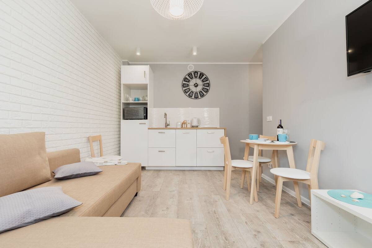 A modern, minimalist living room with a sofa, a dining table with chairs, a kitchen area with cabinets, and a wall-mounted TV.