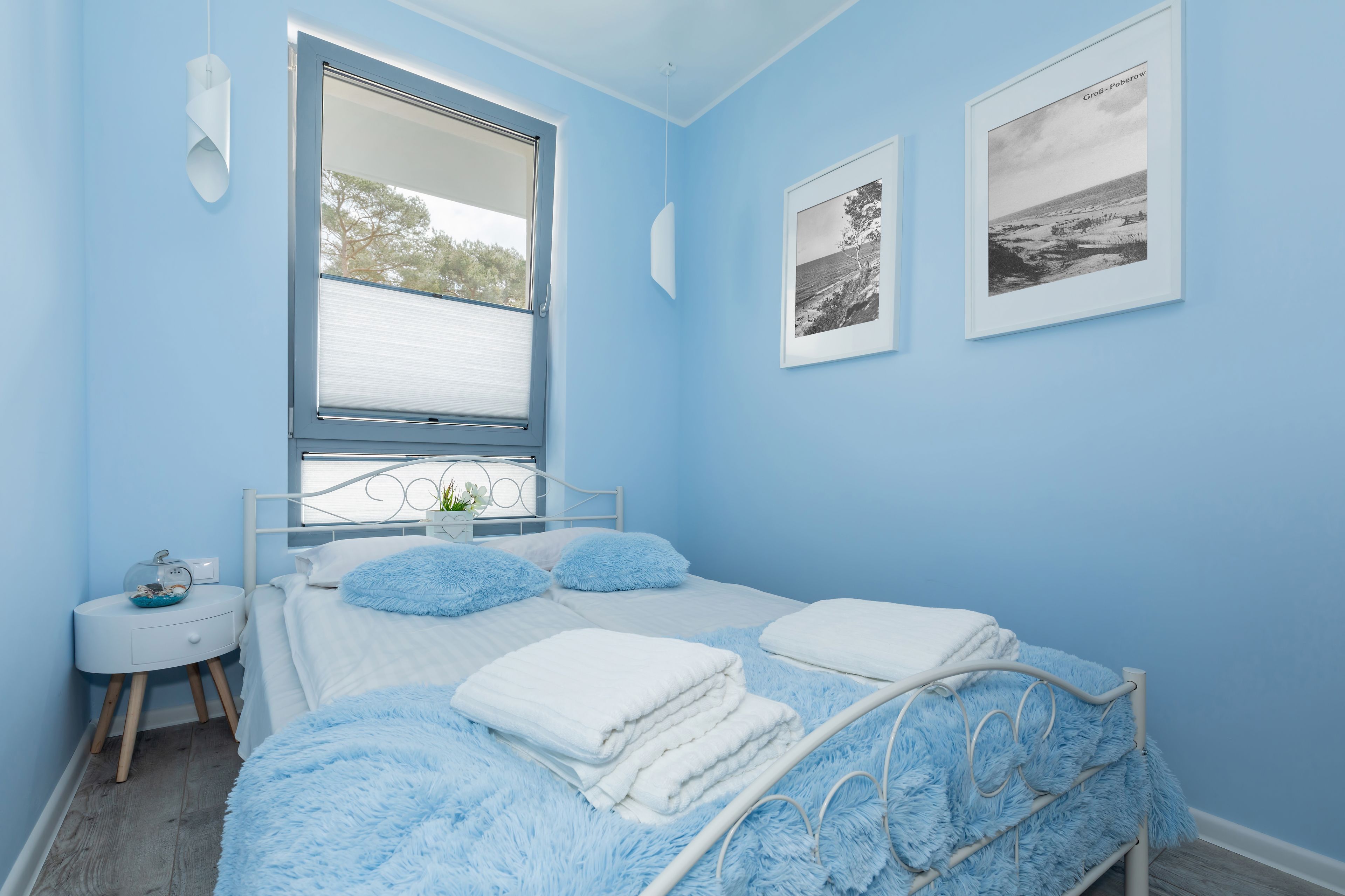 A bedroom with a bed, a window, a bedside table, and two framed pictures on the wall.