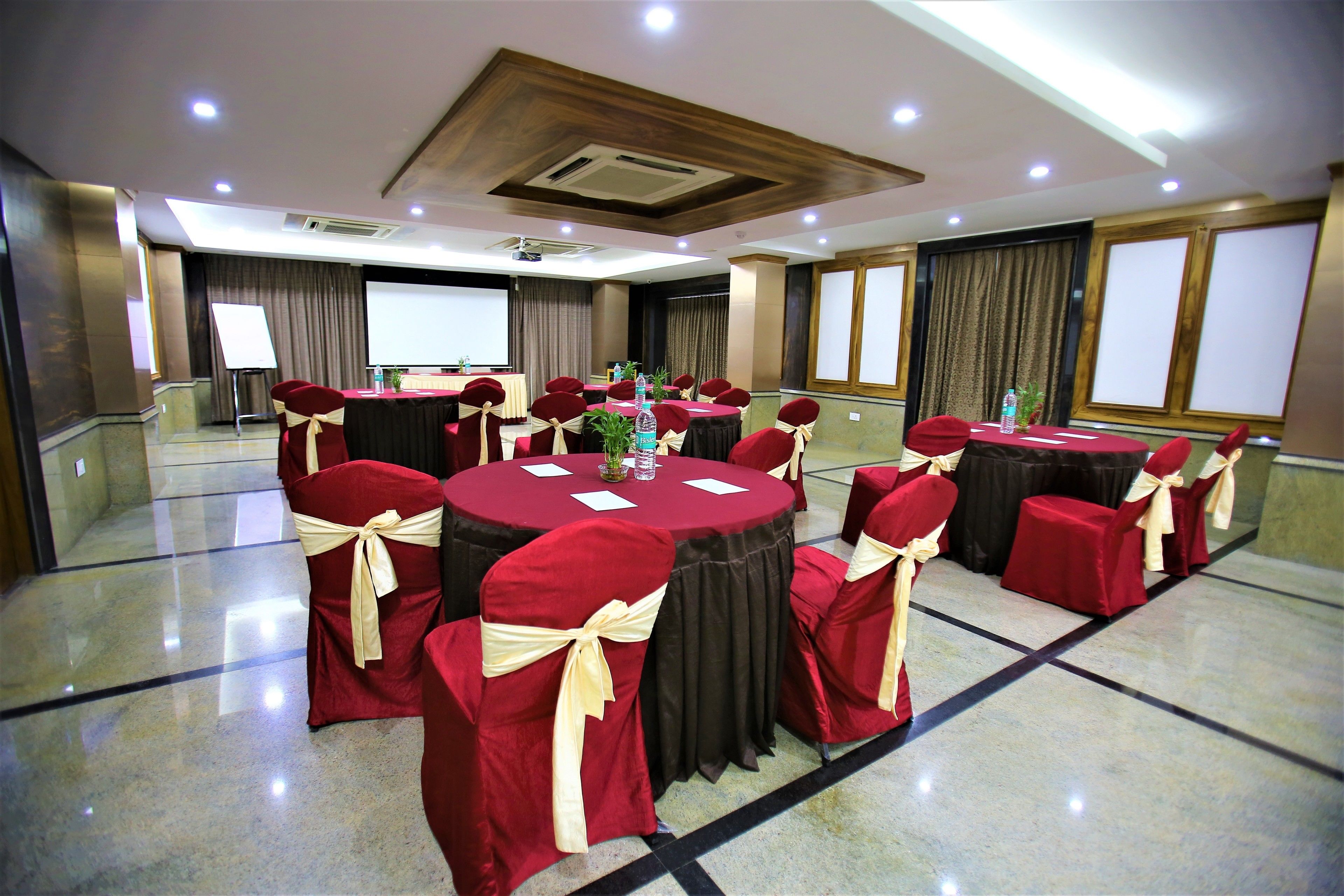 Un salón de conferencias con mesas redondas, sillas rojas con cintas doradas, una pantalla de proyector y un amplio ventanal.