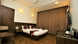 Habitación de hotel con piso de madera, cama con ropa blanca, mesita de noche, escritorio, silla, ventilador de techo y una ventana grande con cortinas.