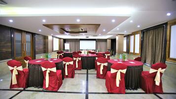 Un salón de conferencias con mesas redondas, sillas rojas con cintas color beige y una pantalla de proyector al frente.