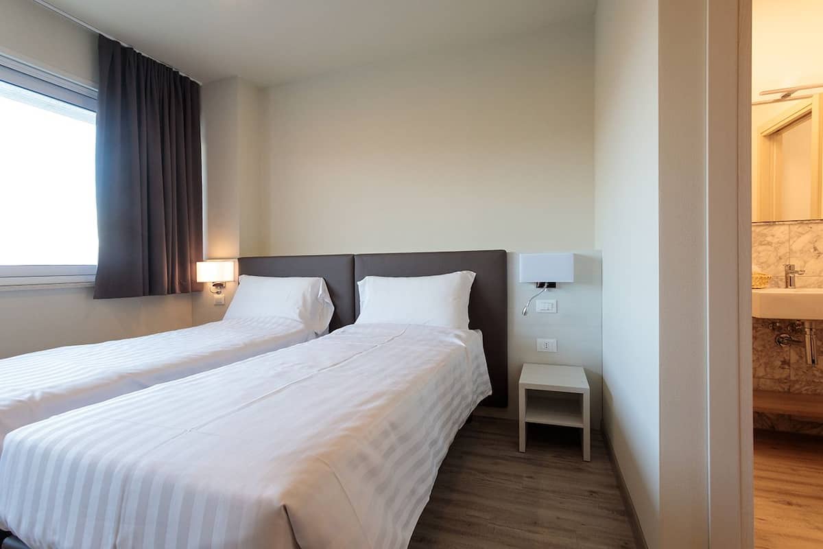 Une chambre d’hôtel avec deux lits simples, une petite table de chevet, une commode et une salle de bain visible à travers une porte ouverte.
