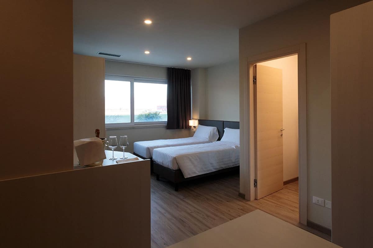 Une chambre d’hôtel avec deux lits, un petit bureau avec une chaise, une fenêtre avec des rideaux et une porte donnant sur une autre pièce.