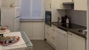 A compact kitchen with white cabinets, a built-in oven, a refrigerator, and a dining table with a clear glass top. - Il Genovino (Genoa)