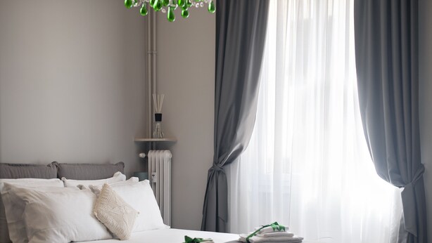 A neatly made bed with white linens and a green towel, grey curtains, and a radiator in a modern hotel room. - Il Genovino (Genoa)