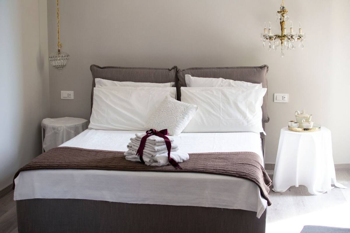 A neatly made bed with white linens, a brown blanket, and a stack of towels with a red ribbon. There is a small round table with a white tablecloth and a tea set on it. A chandelier hangs from the ceiling.