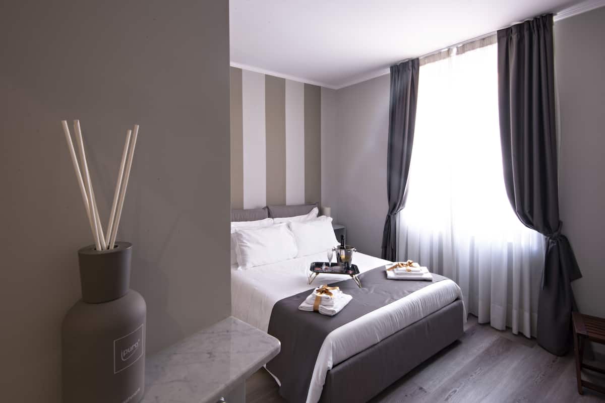 A modern hotel room with a bed, a bedside table, a vase with reed sticks, and a window with curtains.