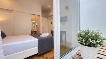 Quarto de hotel moderno com cama, uma pequena mesa de trabalho, uma planta em vaso e uma porta de vidro deslizante para uma varanda.