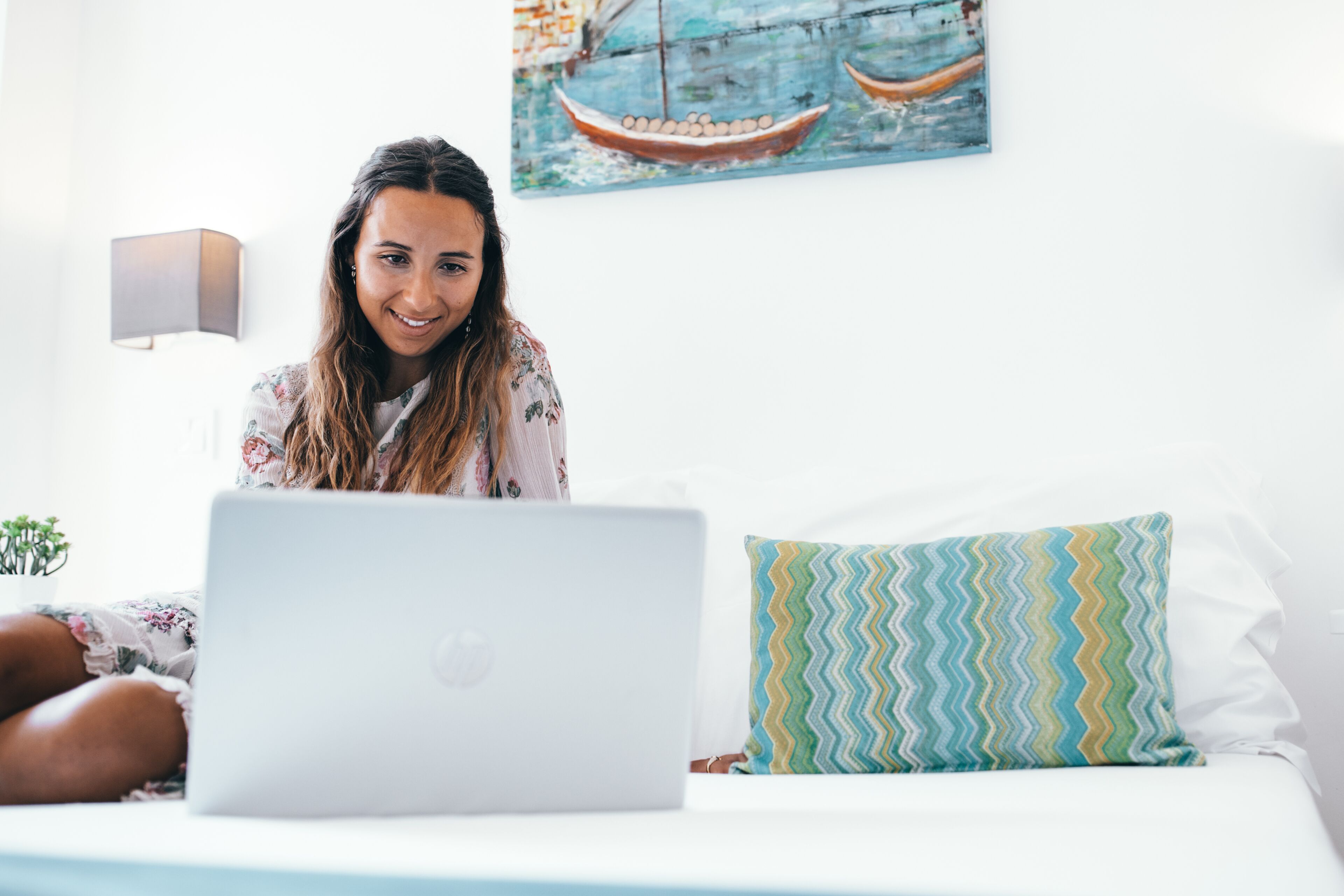 Iemand zit op een bed met een laptop, een sierkussen en een wandlamp.