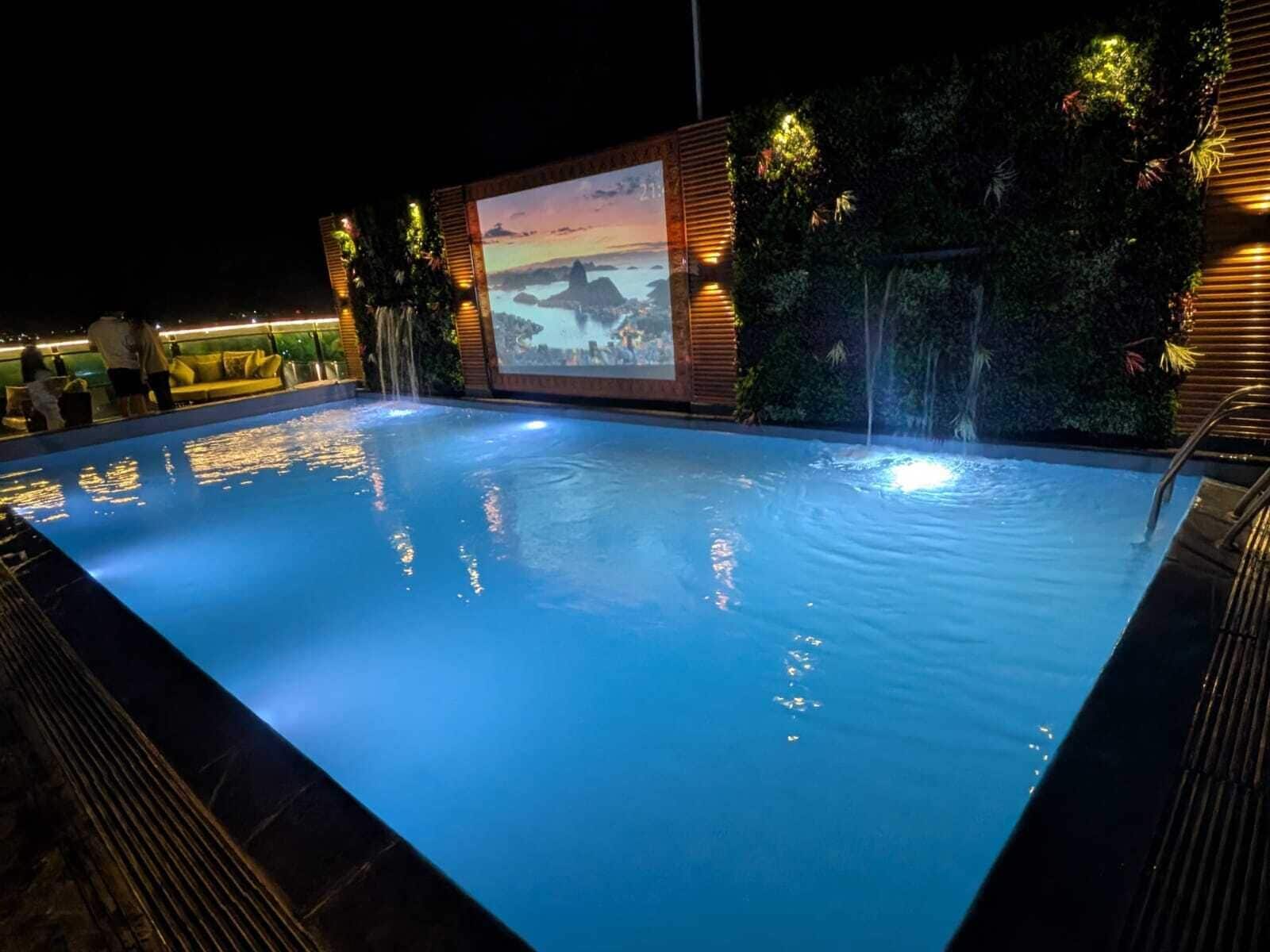 A swimming pool with a waterfall feature, surrounded by a wooden deck and a large screen displaying a scenic view.