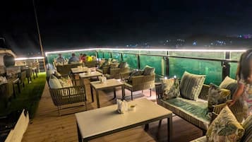 An outdoor deck with seating and tables, a grill, and a view of the city at night.